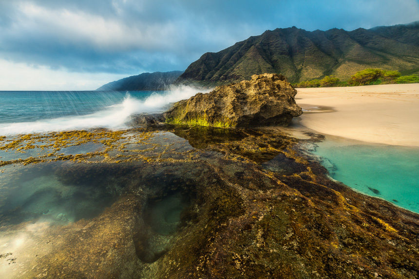 A tropical photo on Oahu of turquoise water tide pools with a wave crashing against a rock.