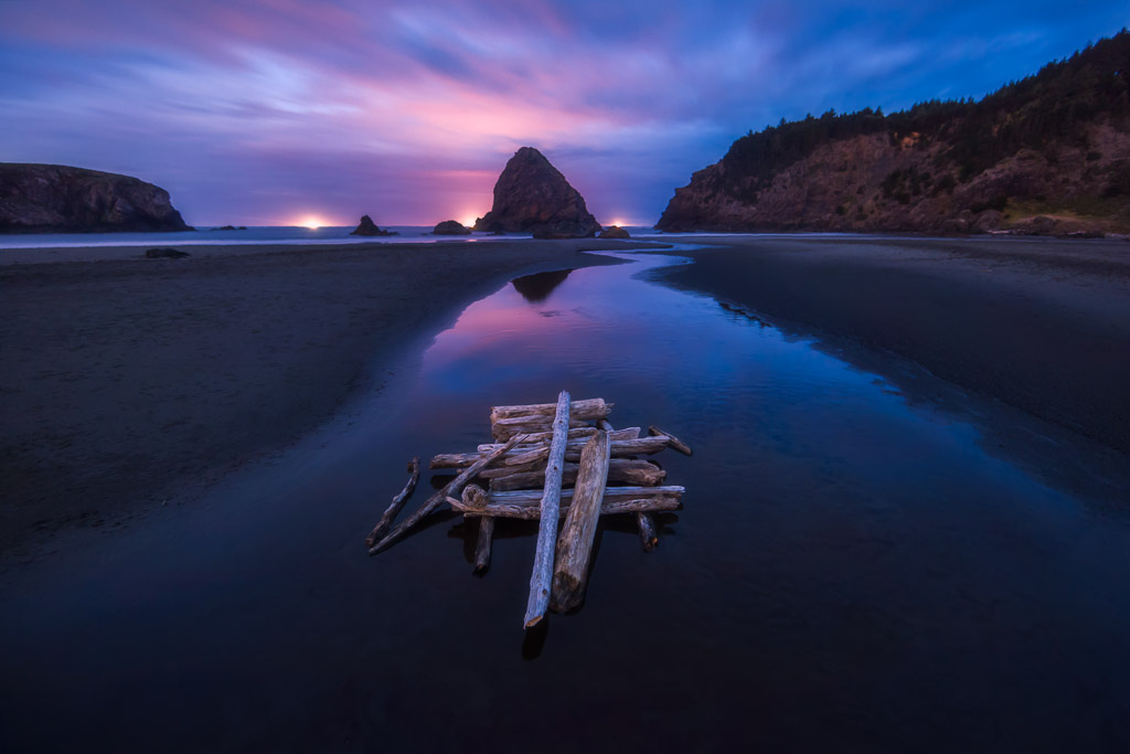 Photo of a stacked logs and a river flowing through a beach towards a sea stack along the oregon coast at twilight